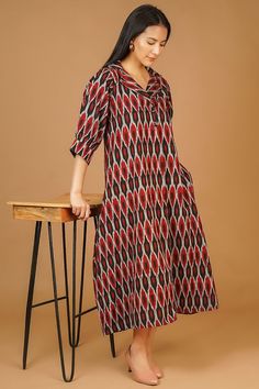 a woman standing next to a wooden table wearing a red and black dress with geometric designs