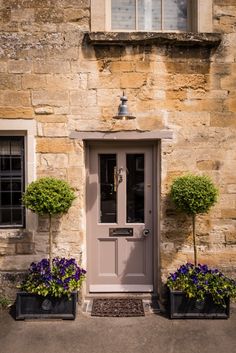 two planters with purple flowers are in front of a stone building and the door is open