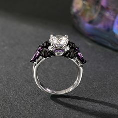 A cool black bat ring boasts a central, dazzling round gemstone, with bat images sculpted on both sides, infusing a touch of the macabre and the enchanting. Surrounding the main stone are sparkling purple and white stones, creating a striking and elegant contrast. Crafted from polished silver metal, the two tone fashion black ring features intricately carved black bat wing decorations, thereby enhancing its mystical charm. Inside, a spider's web-like hollowed-out lining lends an air of whimsy. It's a perfect fusion of sophistication, mystery, and allure, making it a standout accessory for any occasion.Carat Weight: 3.5 ctStone Size: 8 mmStone Type: Jeulia® StoneNumber of Stones: 1 Stone Color: Diamond WhiteStone Shape: RoundCarat Weight: 2.254 ctStone Size: 1.5,1,5*7 mmStone Type: Jeulia® Black Fantasy Rings As Gift, Black Fantasy Style Rings As Gift, Fantasy Style Black Ring For Gift, Elegant Black Skull Ring For Halloween, Elegant Halloween Ring Jewelry, Elegant Halloween Jewelry Rings, Bat Images, Bat Ring, White Stones