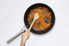 a person holding a wooden spoon over a frying pan filled with shredded carrots