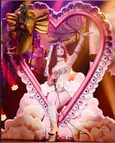 a woman in white dress sitting on top of a pink heart shaped stage with angel wings