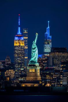 the statue of liberty lit up at night in new york city, with other tall buildings behind it