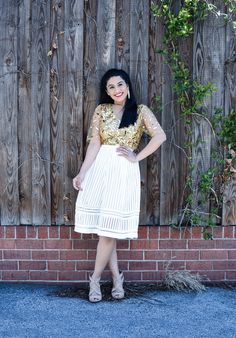 a woman standing in front of a wooden fence wearing a white skirt and gold top