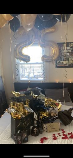 balloons and gifts on a bed in a room with white sheets, black pillows and red rose petals