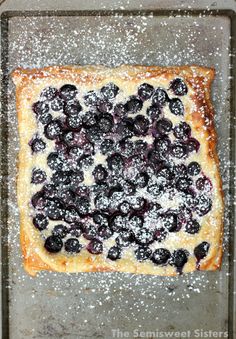 a square pastry topped with blueberries and powdered sugar on a baking sheet covered in powdered sugar