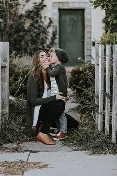 a mother kissing her son while sitting on the ground in front of a wooden fence