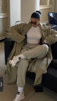 a woman sitting on top of a black couch wearing white sneakers and a trench coat