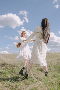 two women in white dresses holding hands and walking through tall grass on a sunny day