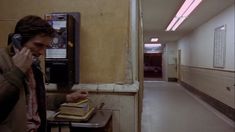 a man standing in a hallway next to a phone and books on a table with a lamp above it