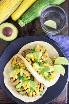 two tacos with corn and avocado on a black plate next to some limes