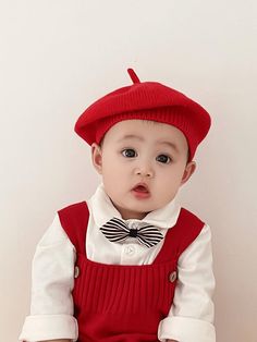 a little boy wearing a red hat and suspenders sitting on the floor in front of a white wall