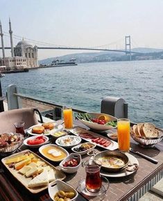 an outdoor dining table with food and drinks on it near the water's edge