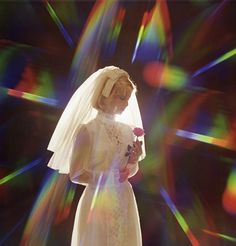 a woman in a wedding dress and veil holding a rose standing against a colorful background