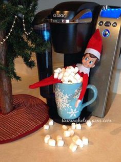 an elf is sitting in a cup of marshmallows next to a coffee maker