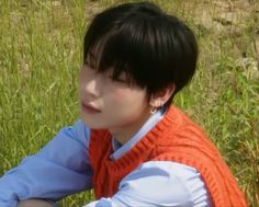 a young man sitting in the grass wearing an orange knitted vest and blue shirt