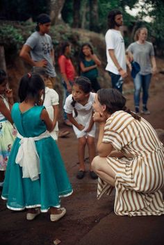 a woman kneeling down next to a group of children on a dirt road with trees in the background