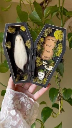 a hand holding an open box with a small white ghost in it and some flowers on the inside