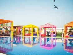 people are sitting at the edge of a swimming pool with brightly colored pavilions in the background