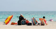 three people in wheelchairs sitting on the beach with an umbrella over their heads, and one person in a wheel chair looking out at the ocean