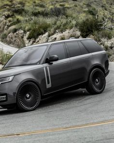 an suv is driving down the road in front of some hills and bushes with trees