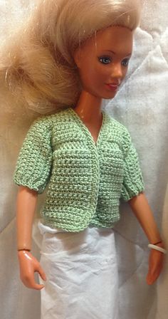 a doll with blonde hair wearing a green shirt and white skirt, standing on a white sheet