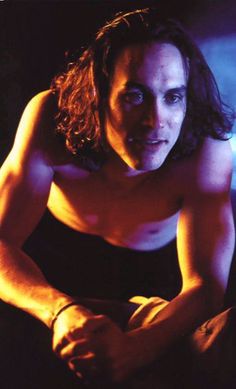 a man with long hair and no shirt is sitting on the floor looking at the camera