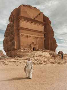 a person walking in front of an ancient building with large rocks on the ground and sky above