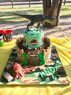 a dinosaur birthday cake on top of a table with dirt around it and trees in the background