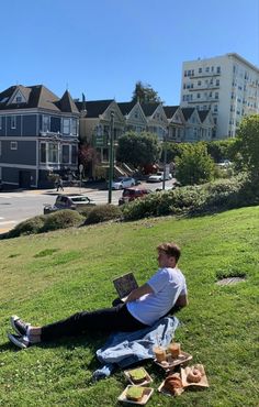 a man sitting on the grass with his laptop in front of him and buildings behind him