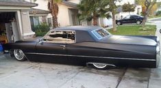 an old black car parked in front of a house next to a driveway and palm trees