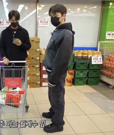 two men standing in front of a shopping cart wearing face masks while looking at their cell phones