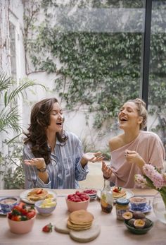 two women sitting at a table with plates of food in front of them and laughing