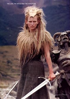 a woman with long hair holding two swords in front of her face and mountains behind her