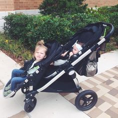 two young children sitting in strollers on the sidewalk next to bushes and shrubbery