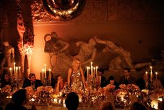 a group of people sitting around a table with candles in front of them and statues behind them