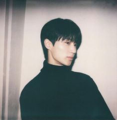 a young man with black hair wearing a black turtle neck shirt and standing in front of a white wall