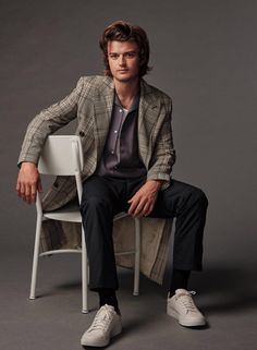 a man sitting on top of a white chair next to a gray wall and wearing a jacket