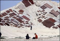 three people sitting on the ground in front of a large rock formation, with one person standing near by