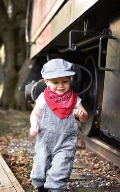 a small child wearing overalls and a hat is standing in front of a train