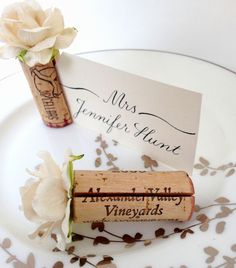 two wine corks are sitting on a plate next to a white rose and a place card