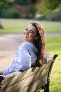 a woman sitting on top of a wooden bench in a park next to a tree