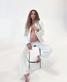 a pregnant woman sitting on top of a stool wearing white pants and a blazer