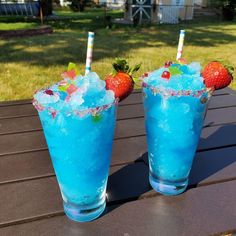 two glasses filled with blue liquid and strawberries on top of a wooden table outside