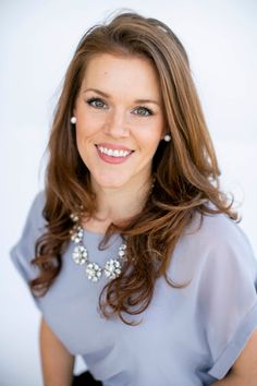 a woman with long brown hair wearing a blue shirt and pearls necklace smiling at the camera