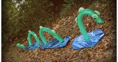 two green plastic birds sitting on top of leaves in the grass next to some trees