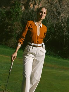 a woman walking across a lush green field holding a golf club and wearing an orange shirt