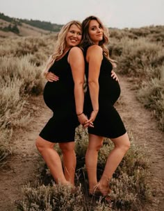 two pregnant women in black dresses posing for a photo on a dirt path with grass and bushes behind them