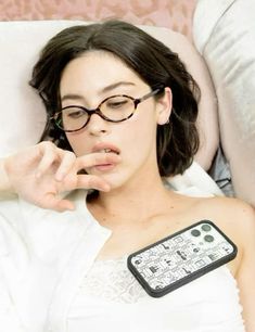 a woman laying in bed wearing glasses and holding a remote control to her ear while looking at the camera