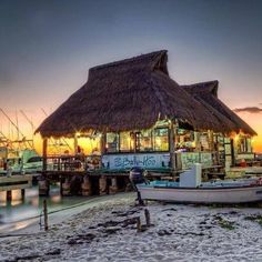 a boat sitting on top of a sandy beach next to a pier at sunset with a tiki hut in the background