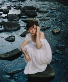 a woman sitting on top of a rock next to a river while talking on a cell phone